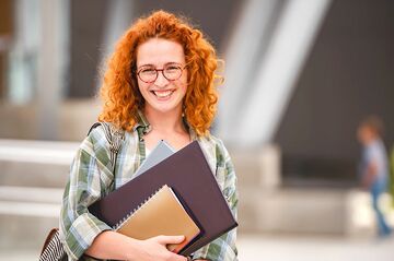 rothaarige Frau mit Brille steht vor einem Gebäude und lächelt. In der Hand Studienunterlagen.