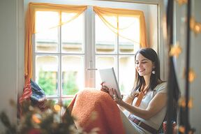 Frau sitzt zuhause am Fenster mit dicker Decke und Studienmaterialien
