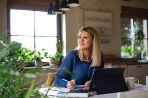Eine blonde Frau sitzt vor ihrem Laptop, macht sich Notizen und schaut verträumt zur Seite.