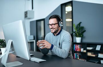 bärtiger Mann mit Headset und Kaffeetasse sitzend vor Bildschirm