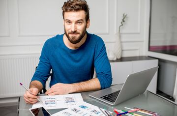 Mann mit blauem Pullover an einem Schreibtisch vor einem Laptop sitzend. 