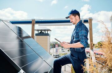 Mann in Blaumann steht auf Dach vor Solar-Panel mit Tablet in der Hand
