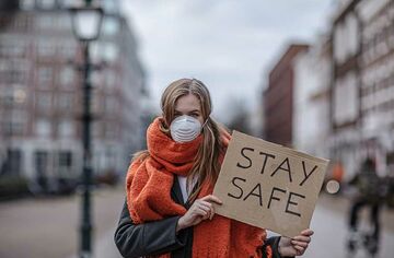 Blonde Frau mit rotem Schal steht draußen mit einem Pappschild auf dem Stay safe steht. 