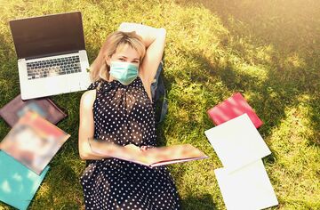 Blonde Frau mit Mundschutz liegt mit Notebook auf Wiese, umgeben von Studienunterlagen