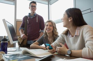 zwei junge Frauen und ein junger Mann sitzen und stehen um einen Schreibtisch