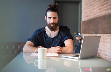 Junger Mann mit Vollbart an Tisch mit Notebook, Notizblock und Tasse