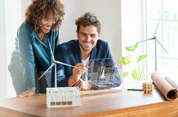 Frau und Mann vor Schreibtisch mit transparentem Bildschirm und Modellen von Windrädern und Gebäuden.