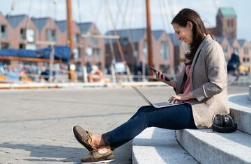 Junge Frau sitzt auf Treppenstufen am Hafen mit Notebook und Smartphone