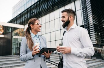 Frau und Mann stehen auf Treppe, unterhalten sich und halten Notebook und Becher in der Hand