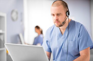 Männliche Pflegekraft sitzt mit Headset vor Notebook.