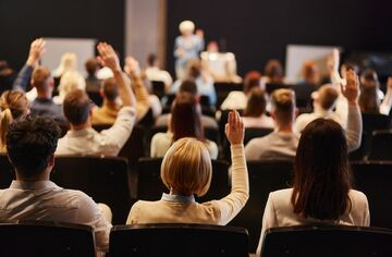 Viele Menschen in Rückansicht sitzen auf einer Internationalen Konferenz und hören einen Vortrag. Einige heben die Hände, um Fragen zu stellen.