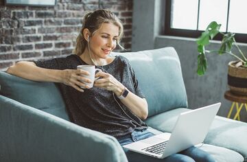 Frau mit Kaffeetasse in Hand und Laptop auf Schoß sitzt auf Sofa