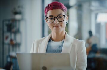 Eine Person hält einen geöffneten Laptop und schaut lächelnd in die Kamera.