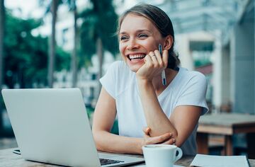 Junge Frau sitzt draußen in Street Cafe mit Notebook am Tisch und lächelt.