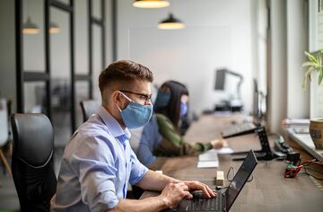 Zwei Personen mit Corona-Masken sitzen an Schreibtisch und arbeiten mit Notebooks