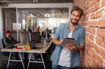 Junger Mann mit Tablet in der Hand lehnt an Ziegelwand in einem modernen Büro und lächelt in Kamere.