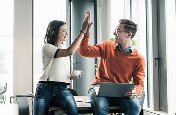 Junge Frau und Mann sitzen auf Tisch, freuen sich und geben High Five.