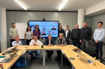 Project Labs in Linz Menschen sitzen und stehen um einen Tisch herum und lächeln in die Kamera. Im Hintergrund ist ein großer Bildschirm, auf dem weitere Menschen iun einer digitalen Konferenz zugeschaltet sind.
