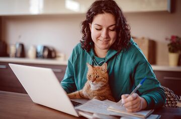 Dunkelhaarige Frau an Schreibtisch mit Notebook macht Notizen auf Papier und hat eine Katze auf dem Schoß.