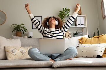 Eine Person mit Kopfhörern und Laptop auf dem Schoß sitzt auf Sofa und reckt beide Arme jubelnd nach oben.