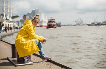 Blonde Frau im gelben Regenmantel sitzt an den Landungsbrücken am Wasser und blickt in die Kamera; im Hintergrund Schiffe und die Elbphilharmonie.