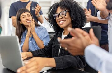 junge Frau mit Brille sitzt mit zufriedenem Blick vor Laptop und erhält Applaus von umstehenden Personen