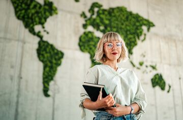 Junge Frau mit blonden Haaren und Brille steht mit Notebook in der Hand vor einer Wand mit einer moosbedeckten grünen Weltkarte