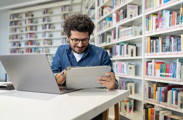 Lockiger Mann sitzt in an Schreibtisch vor Bücherregalen und hantiert mit Laptop und Tablet.