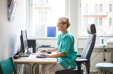 Kurzhaarige Pflegebeschäftigte in Büro an Schreibtisch vor Computer.