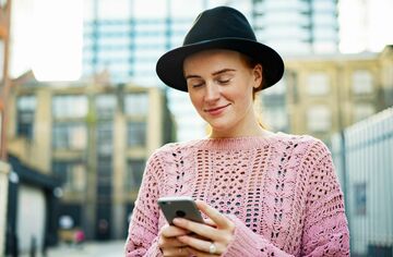 Junge Frau mit Hut blickt auf Smartphone und lächelt