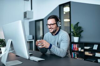 bärtiger Mann mit Headset und Kaffeetasse sitzend vor Bildschirm 