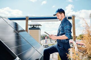 Mann in Blaumann steht auf Dach vor Solar-Panel mit Tablet in der Hand