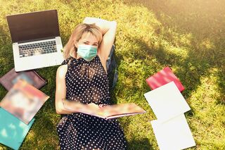 Blonde Frau mit Mundschutz liegt mit Notebook auf Wiese, umgeben von Studienunterlagen