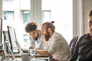 Zwei junge Männer sitzen vor Notebooks und diskutieren