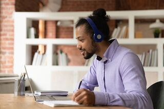 Ein junger Mann sitzt mit Headset vor einem Laptop und macht sich Notizen.