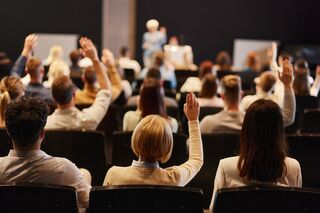 Viele Menschen in Rückansicht sitzen auf einer Internationalen Konferenz und hören einen Vortrag. Einige heben die Hände, um Fragen zu stellen.