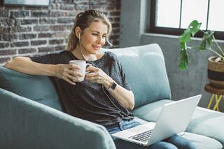 Frau mit Kaffeetasse in Hand und Laptop auf Schoß sitzt auf Sofa