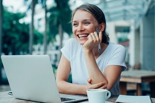 Junge Frau sitzt draußen in Street Cafe mit Notebook am Tisch und lächelt.