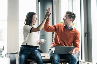 Junge Frau und Mann sitzen auf Tisch, freuen sich und geben High Five.