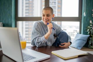 Eine junge Person mit rasierten Haaren sitzt an einem Schreibtisch und schaut lächelnd auf einen Laptop. Sie hält einen Stift in der Hand und hat ein aufgeschlagenes Notizbuch vor sich.