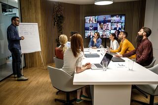 Lernsituation in einem Lehrraum mit Personen an Tisch und zugeschalteten Online-Teilnehmenden auf großem Screen.