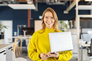 Eine Frau mit Laptop im Arm lächelt in die Kamera.