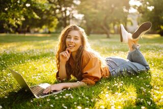 Junge Frau liegt auf einer Wiese, arbeitet lächelnd am Laptop – entspanntes Lernen im Freien bei Sonnenschein.