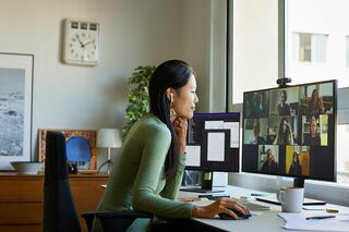 Junge Frau sitzt an Schreibtisch vor großem Monitor, auf dem andere Menschen zu sehen sind, und nimmt an einer Online-Lehrveranstaltung teil. 