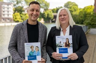 Prof. Dr. Michael Heinrich-Zehm steht links neben Prof. Dr. Eske Jung vor dem Osterbekkanal in Hamburg; beide halten je einen Studienführer der HFH in der Hand.