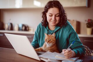 Dunkelhaarige Frau an Schreibtisch mit Notebook macht Notizen auf Papier und hat eine Katze auf dem Schoß.