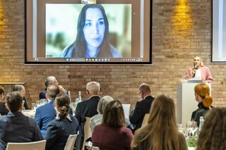 Blick in die Veranstaltung zur Verleihung des Studienpreises 2025 mit sitzenden Gästen, einer Rednerin an Sprechpult und der Preisträgerin auf einem Screen an der Wand.