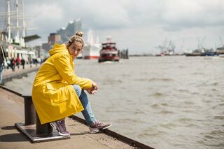 Blonde Frau im gelben Regenmantel sitzt an den Landungsbrücken am Wasser und blickt in die Kamera; im Hintergrund Schiffe und die Elbphilharmonie.