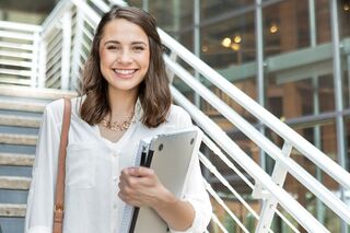 Junge Frau mit Unterlagen und Notebook in der Hand steht auf einer Treppe und lächelt in die Kamera.