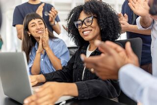 junge Frau mit Brille sitzt mit zufriedenem Blick vor Laptop und erhält Applaus von umstehenden Personen